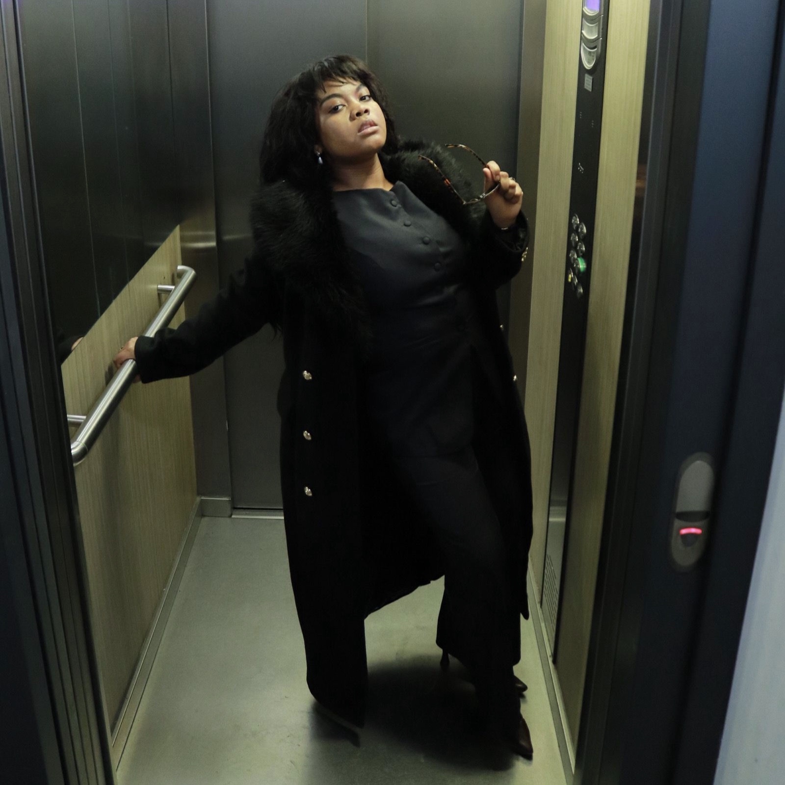 Rosa Zand en long manteau noir à col de fourrure dans un ascenseur