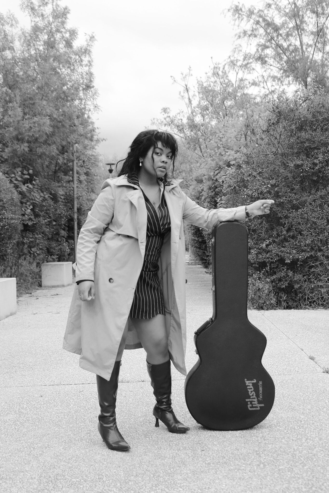 Rosa Zand en trench et robe rayée, appuyée sur un étui de guitare Gibson, photo noir et blanc